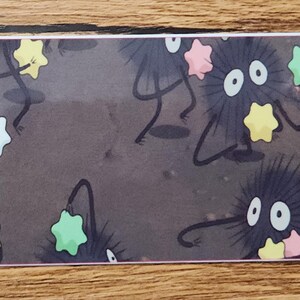 Peut inclure: Un marque-page rectangulaire au fond gris fonc&eacute; orn&eacute; de plusieurs petites cr&eacute;atures rondes, noires et duveteuses aux grands yeux blancs. Ces cr&eacute;atures tiennent de petits bonbons en forme d'&eacute;toile dans des tons jaune, rose et vert clair.