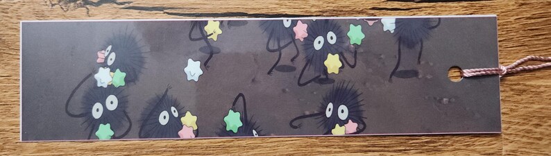 Peut inclure: Un marque-page rectangulaire au fond gris fonc&eacute; orn&eacute; de plusieurs petites cr&eacute;atures rondes, noires et duveteuses aux grands yeux blancs. Ces cr&eacute;atures tiennent de petits bonbons en forme d'&eacute;toile dans des tons jaune, rose et vert clair.