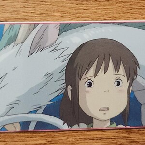 Peut inclure: Un marque-page rectangulaire en papier sur fond bleu illustrant une jeune personne aux cheveux bruns, un grand dragon blanc, une petite cr&eacute;ature grise ressemblant &agrave; une souris et un minuscule esprit de suie vert, avec un cordon bleu sarcelle.