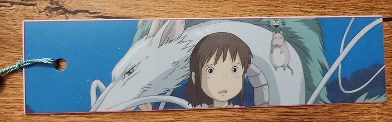 Peut inclure: Un marque-page rectangulaire en papier sur fond bleu illustrant une jeune personne aux cheveux bruns, un grand dragon blanc, une petite cr&eacute;ature grise ressemblant &agrave; une souris et un minuscule esprit de suie vert, avec un cordon bleu sarcelle.