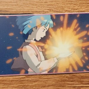 Peut inclure: Un marque-page rectangulaire sur fond bleu fonc&eacute; avec l'illustration imprim&eacute;e d'un personnage aux cheveux courts bleus tenant une source de lumi&egrave;re jaune vif &eacute;clatante qui &eacute;met des &eacute;tincelles. Un petit trou est perc&eacute; &agrave; gauche avec un cordon jaune.