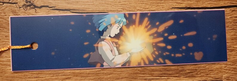 Peut inclure: Un marque-page rectangulaire sur fond bleu fonc&eacute; avec l'illustration imprim&eacute;e d'un personnage aux cheveux courts bleus tenant une source de lumi&egrave;re jaune vif &eacute;clatante qui &eacute;met des &eacute;tincelles. Un petit trou est perc&eacute; &agrave; gauche avec un cordon jaune.