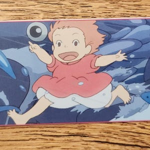 Peut inclure: Un marque-page rectangulaire en papier pr&eacute;sentant une illustration color&eacute;e d'un petit enfant aux cheveux p&ecirc;che portant une robe rose, courant sur des vagues bleu fonc&eacute; entour&eacute; de plusieurs poissons bleus et d'une grande cr&eacute;ature marine.