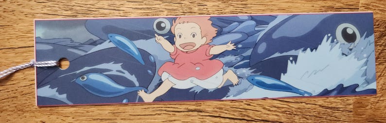 Peut inclure: Un marque-page rectangulaire en papier pr&eacute;sentant une illustration color&eacute;e d'un petit enfant aux cheveux p&ecirc;che portant une robe rose, courant sur des vagues bleu fonc&eacute; entour&eacute; de plusieurs poissons bleus et d'une grande cr&eacute;ature marine.