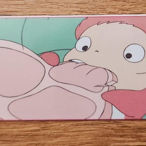 Peut inclure: Un marque-page rectangulaire en papier pr&eacute;sentant une illustration color&eacute;e d'un petit personnage aux cheveux roses et aux grands yeux mordant dans un gros morceau de viande rose. Le marque-page poss&egrave;de un pompon vert attach&eacute; par un trou.
