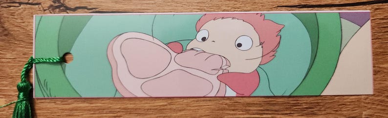 Peut inclure: Un marque-page rectangulaire en papier pr&eacute;sentant une illustration color&eacute;e d'un petit personnage aux cheveux roses et aux grands yeux mordant dans un gros morceau de viande rose. Le marque-page poss&egrave;de un pompon vert attach&eacute; par un trou.