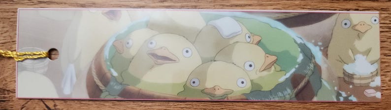 Peut inclure: Un marque-page rectangulaire en papier illustr&eacute; de plusieurs cr&eacute;atures rondes et jaune p&acirc;le, semblables &agrave; des oiseaux, se baignant ensemble dans une grande cuve en bois. Un petit trou est perc&eacute; &agrave; gauche avec un fin cordon dor&eacute; m&eacute;tallique.