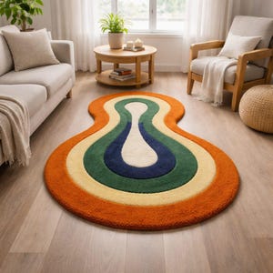 Puede incluir: Una alfombra colorida de estilo retro con una forma orgánica única. La alfombra presenta bandas concéntricas de color naranja, amarillo, verde, azul marino y blanco. Está colocada sobre un suelo de madera clara, añadiendo un acento vibrante a la decoración de la habitación.
