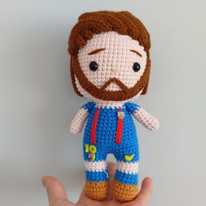 May include: A handmade crochet doll of a soccer player, featuring a brown beard and hair, wearing a blue uniform with red suspenders and the number 10. The doll has a light skin tone and is holding a soccer ball.