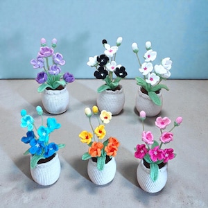 Puede incluir: Seis arreglos florales de ganchillo hechos a mano en macetas blancas pequeñas. Las flores son de varios colores, incluyendo morado, negro, blanco, azul, amarillo, naranja y rosa. Las macetas tienen un aspecto texturizado y tejido, y las flores tienen hojas verdes.