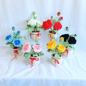 Puede incluir: Una colección de plantas de rosas hechas a mano con ganchillo en varios colores, incluyendo azul, rosa, rojo, amarillo, blanco y negro. Cada rosa está en una pequeña maceta blanca de ganchillo con una cinta roja. Las rosas tienen hojas verdes.