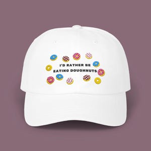 May include: White baseball cap with embroidered donuts in various colors. The cap features the text "I'D RATHER BE EATING DOUGHNUTS" in black. The donuts are yellow, pink, blue, and brown with different toppings. The cap has a curved brim and a rounded crown.