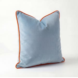 Könnte beinhalten: Ein quadratisches, hellblaues Kissen mit einer weichen, samtigen Textur. Das Kissen hat einen leuchtend orangefarbenen Rand, der einen auffälligen Kontrast erzeugt. Dieser Artikel ist ein dekoratives Element, geeignet für ein Bett oder Sofa.
