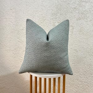 Könnte beinhalten: Ein quadratisches, hellgrünes Zierkissen mit Fischgrätenmuster. Das Kissen besteht aus einem strukturierten Stoff und wird auf einem kleinen, weißen Tisch mit Holzbeinen präsentiert. Die Farbe des Kissens ist ein gedämpftes, weiches Grün.