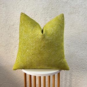 May include: A square decorative pillow in a vibrant chartreuse green color. The pillow features a textured chevron pattern in a lighter shade of green. The pillow is placed on a small, white, round table with wooden legs.