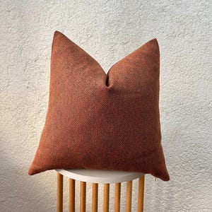 May include: A square, rust-colored pillow with a woven herringbone pattern. The pillow is placed on a small, white, round table with wooden legs. The pillow's texture and color create a warm, inviting aesthetic.