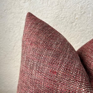 Peut inclure: Gros plan sur un coussin texturé de couleur bordeaux. Le tissu a un aspect tissé avec un mélange de fils rouges et gris. Le coin du coussin est visible, mettant en valeur le tissage et les variations de couleur du tissu. C'est un article de décoration intérieure.