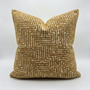 May include: A square decorative pillow with a geometric pattern. The pillow features a textured, golden-brown fabric with a white, maze-like design. The pillow is a neutral color and would complement a variety of home decor styles.