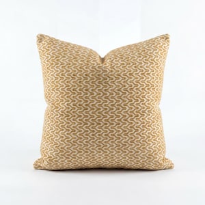 May include: A square decorative pillow with a repeating geometric pattern in shades of gold and cream. The pillow has a soft, textured appearance and is likely made of fabric. The design is symmetrical and covers the entire surface.