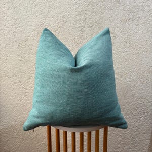 May include: A teal cushion with a textured weave, placed on a small, round, white table with wooden legs. The cushion's colour is a muted teal, and it appears soft. Ideal for adding a touch of colour to a room.