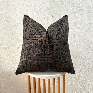 Può includere: Un cuscino quadrato decorativo con un motivo strutturato nero e marrone. Il cuscino è posto su un tavolino rotondo bianco con gambe in legno. Il design del cuscino presenta un motivo a griglia intrecciata.