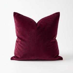 Op de afbeelding: Een vierkant sierkussen met een hoes van diepbordeauxrode fluwelen stof. Het kussen heeft een zachte, pluche textuur met een consistente donkerrode kleur over het gehele oppervlak, afgewerkt met strakke randen en een volle, gevulde vorm.