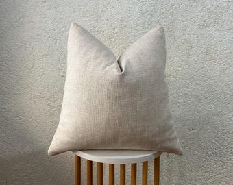 May include: A beige square pillow with a textured fabric, resting on a small white and wood side table. The pillow has a simple, neutral design, suitable for home decor.