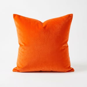 May include: A square, orange velvet pillow. The pillow is a solid, vibrant orange color and has a soft, plush appearance. The pillow is likely used for home decor and adds a pop of color to any room.