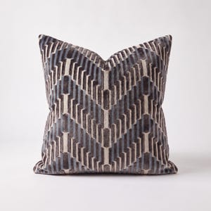 May include: A square throw cushion featuring a textured velvet fabric with a repeating geometric chevron pattern in shades of dark charcoal grey, slate blue, and cream. The design consists of vertical and angled lines creating a structured look.