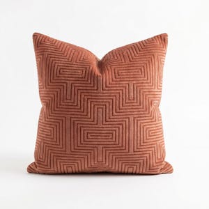 May include: A square throw pillow in a terracotta orange color featuring a raised, textured geometric pattern of interlocking rectangles and lines that create a repeating maze-like design across the entire front surface of the fabric.
