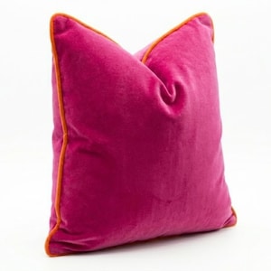 May include: A magenta velvet pillow with orange piping. The square pillow is a vibrant accent piece, adding a pop of color to any room. The plush texture and contrasting trim create a luxurious feel.