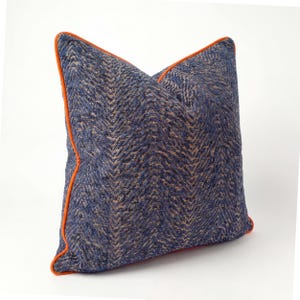 May include: A square decorative pillow with a navy blue and tan herringbone pattern. The pillow has an orange trim around the edges. The pillow is a decorative item for a bed or sofa.