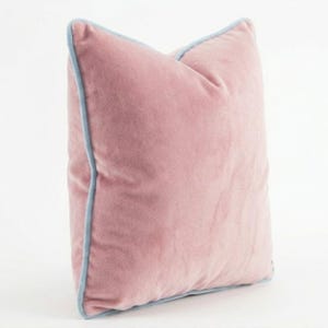 May include: A plush, pink velvet pillow with a light blue trim. The square pillow is a soft, muted rose color and has a subtle sheen. The light blue trim outlines the edges of the pillow, adding a contrasting detail.