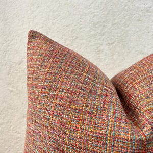 Op de afbeelding: Close-up van twee decoratieve sierkussens. De kussens hebben een textiel, geweven stof in een mix van kleuren, waaronder rood, oranje, geel en blauw. De kussens staan tegen een neutrale achtergrond.