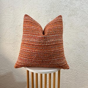 May include: A square decorative pillow with a textured, striped pattern in shades of orange, white, and brown. The pillow is placed on a small, round, white table with wooden legs. The pillow's dimensions are not visible.