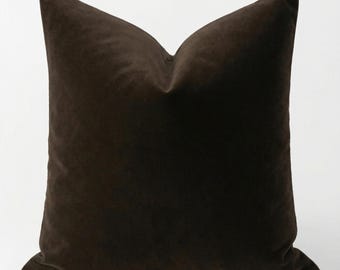 May include: A square, dark brown velvet pillow with a soft, textured appearance. The pillow is a solid color and has a classic, simple design. The pillow is likely used for home decor.