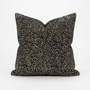 May include: A square cushion with a black and beige speckled pattern. The cushion has a soft, textured appearance and is set against a white background. The cushion is a home decor item.