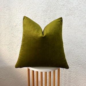 May include: A square olive green velvet pillow. The pillow is placed on a small, round, white table with wooden legs. The background is a textured white wall. The pillow is a solid color and appears soft and plush.