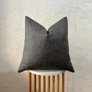 May include: A dark gray square pillow with a textured fabric, resting on a small white and wood side table. The pillow has a simple, modern design and is a neutral color.