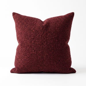 Op de afbeelding: Een vierkant sierkussen in een diepe bordeauxrode kleur met een sterk getextureerd, noppig oppervlak in bouclé-stijl. Het kussen heeft een volle, gevulde uitstraling met duidelijke hoeken en een consistent, dicht weefsel over het gehele oppervlak.