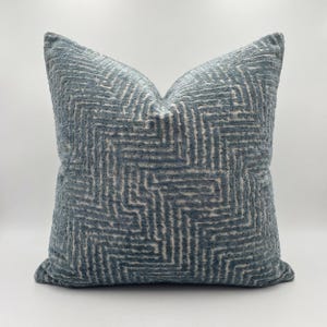 Può includere: Un cuscino decorativo quadrato con un motivo geometrico strutturato nei toni del blu e bianco sporco. Il cuscino ha un aspetto morbido e soffice ed è progettato per l'arredamento della casa. Il motivo è un design a zig-zag ripetuto.
