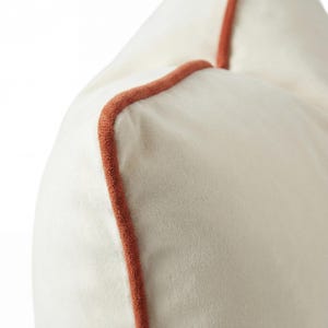 Può includere: Primo piano di un cuscino color crema con bordatura color ruggine. Il cuscino ha una consistenza morbida e vellutata. La bordatura segue i bordi, creando un bordo definito. Il design è semplice ed elegante.