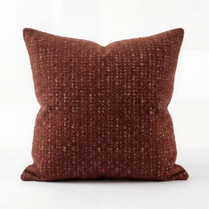 May include: A square decorative pillow in a deep reddish-brown color. The pillow has a textured, woven appearance with small flecks of white throughout. The pillow is a solid color and has a soft, inviting look.