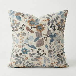 Könnte beinhalten: Quadratisches Zierkissen mit floralem Muster. Das Kissen hat einen cremefarbenen Hintergrund mit einem sich wiederholenden Design aus blauen, braunen und beigen Blumen und Blättern. Das Kissen hat ein weiches, strukturiertes Aussehen.