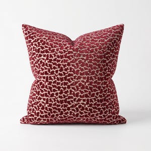 May include: A square throw pillow featuring a deep red velvet fabric with a raised, textured pattern of irregular, interconnected shapes in a lighter cream color. The pillow has a plush, filled appearance with soft, rounded corners and edges.