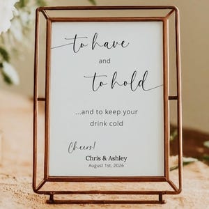 May include: A copper-colored metal framed sign with the text "to have and to hold" in elegant script. The sign also includes the text "...and to keep your drink cold" and "Cheers!" with a date and names. The sign is on a table with floral arrangements.