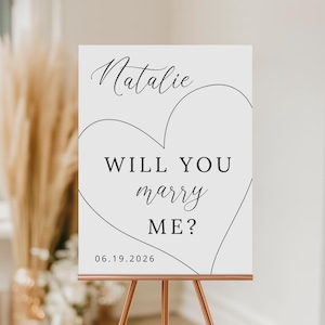 Könnte beinhalten: Ein weißes Schild mit den Worten "Natalie, Will You Marry Me?" in schwarzer Schrift und einer Herzumrandung. Das Datum "06.19.2026" ist unten aufgedruckt. Das Schild wird auf einer kupferfarbenen Staffelei präsentiert, passend für eine Hochzeit oder Verlobung.