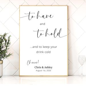 May include: A framed white sign with gold trim features elegant cursive text: "to have and to hold...and to keep your drink cold." Below, it reads "Cheers!" and the names "Chris & Ashley" with a date. A small potted plant and wine glasses are visible.