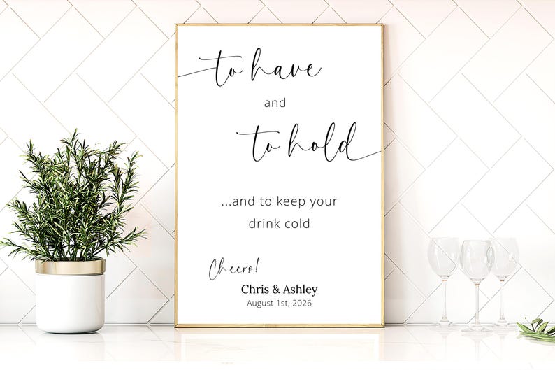 May include: A framed white sign with gold trim features elegant cursive text: "to have and to hold...and to keep your drink cold." Below, it reads "Cheers!" and the names "Chris & Ashley" with a date. A small potted plant and wine glasses are visible.