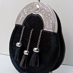 Puede incluir: Un sporran de piel negra con un cierre de metal plateado pulido con intrincados patrones de nudos celtas en relieve. La parte frontal de la bolsa está adornada con tres cadenas plateadas a juego, cada una terminada en una borla de piel negra.